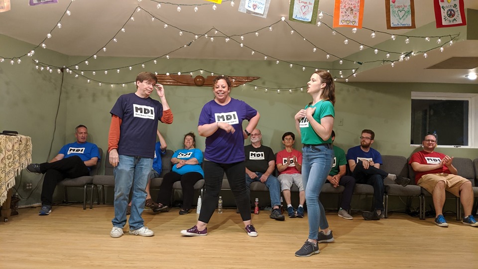 A photo of Steve, Jamie, and Maddie in an improv scene at a show with Scott, Michael, Harris, Jim, Paula, tony, Devon, and Andrew sitting behind them facing the audience. They all wear colorful t-shirts with the MDI logo upon them. A string of cafe lights decorates the ceiling.