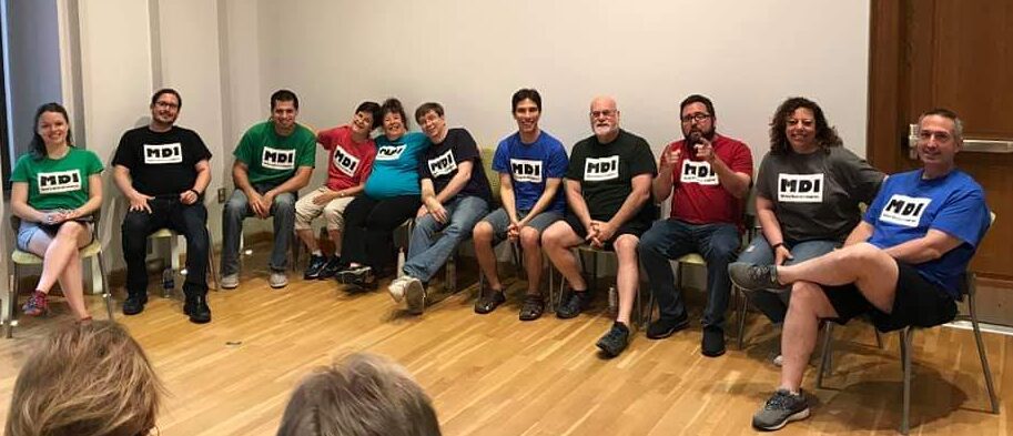 An image of Maddie, Tim, Tony, Paula, Harris, Steve, Michael, Jim, Ted, Jamie, and Scott, sitting in chairs at an improv show, smiling toward where the audience is sitting. They each wear colorful shirts with the MDI logo upon them.