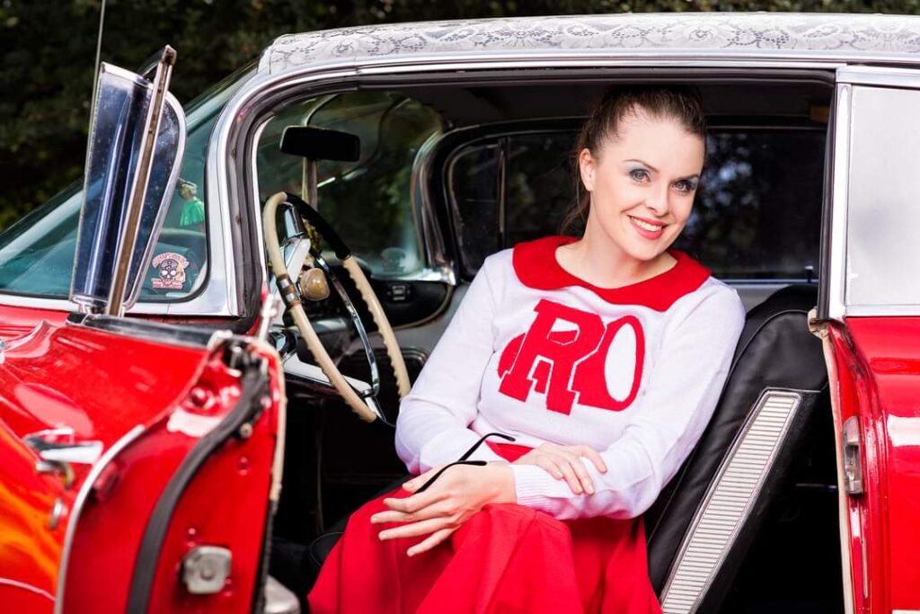 A photo of Maddie dressed in a character costume (as Patti Simcox from the movie Grease), sitting in a red classic car from the 1950s era, wearing an cheerleading costumer from the same period (a white sweater with a red letter R) and a red poodle skirt.