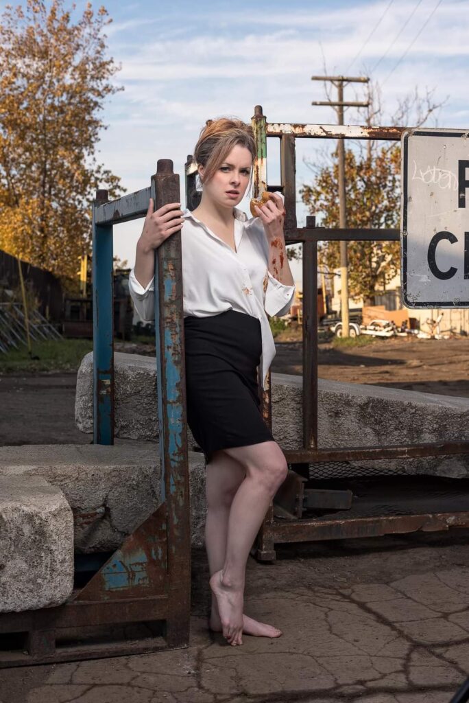 A photo of Maddie in a black pencil skirt and white blouse that is untucked, holding a cheeseburger in her left hand, with ketchup dripping down her left arm, and looking at the camera with attitude.