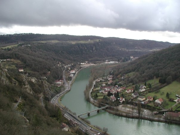 An aerial view of the Doubs river in Besancon, France, with hills on either side and houses along the river. A bridge connects both sides of the river.