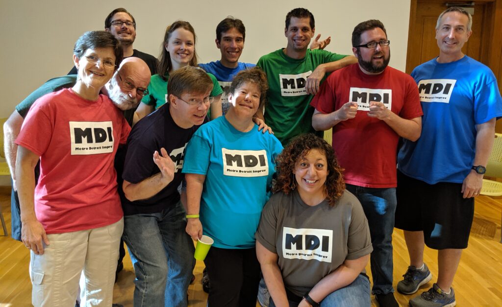 A photo of Tim, Maddie, Michael, Tony, Ted, Scott, Paula, Jim, Steve, Harris, and Jamie posing for the camera, all wearing colorful t-shirts with the MDI logo upon them at an improv show.