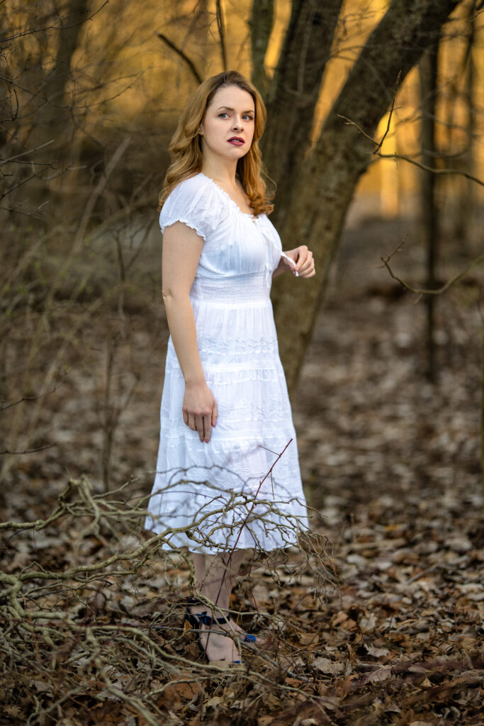 A photo of Maddie in a white dress, standing in the woods during what appears to be a cold season with no leaves on the trees. She is holding a string on her dress with her left hand and gazing off into the distance with a pensive look on her face.