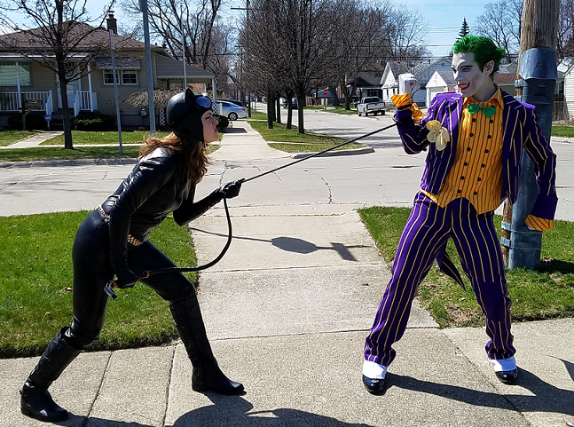 A photo of Maddie dressed as Catwoman, in a full body black leather outfit, tugging on a whip that Michael dressed as Joker is pulling in his direction with a sneer on his face. They are standing on a neighborhood block corner, with houses and cars in the background.