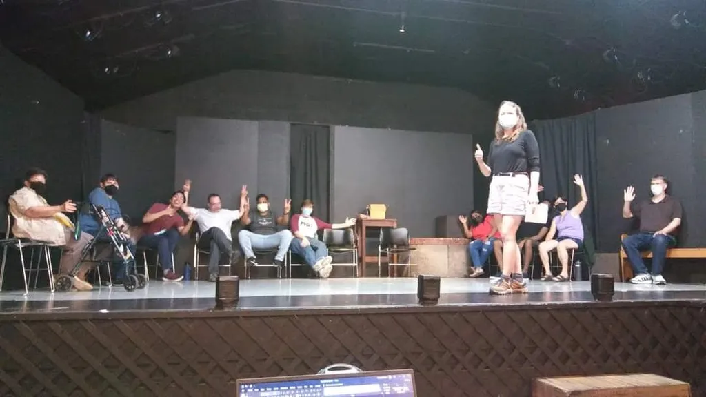 A photo of Maddie with a COVID mask on her face, giving a thumbs up, standing on a stage with ten improvisers sitting and waving on chairs in the background.