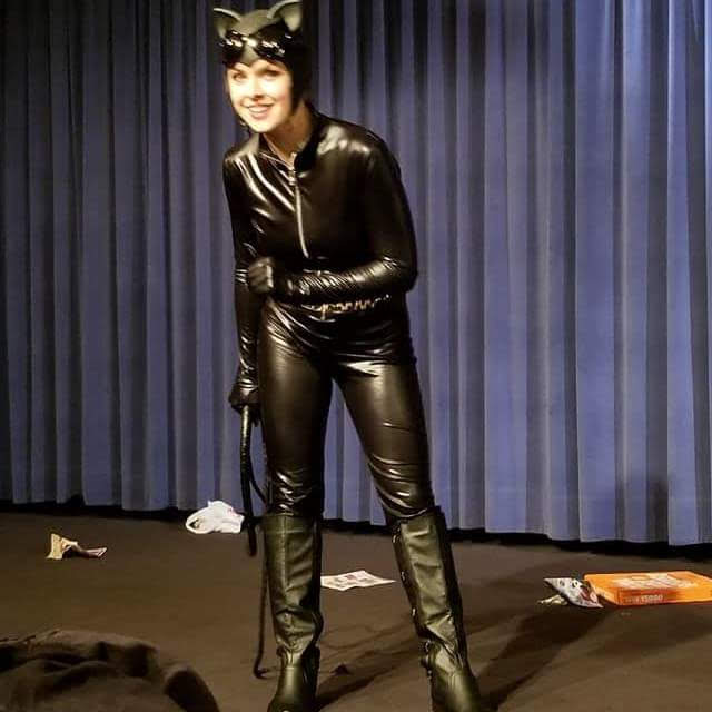 A full body shot of Maddie in her improv show dressed as Catwoman, holding a leather whip, and looking out at the audience with a smile on her face