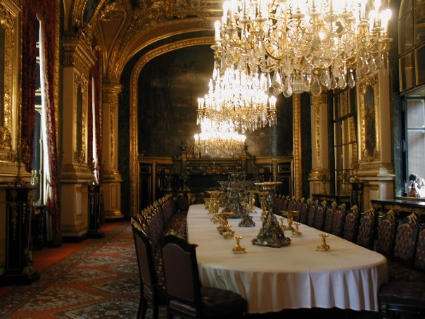 A photo of a long white table surrounded by many fancy chairs with ornate chandeliers hovering overhead from a museum in France.