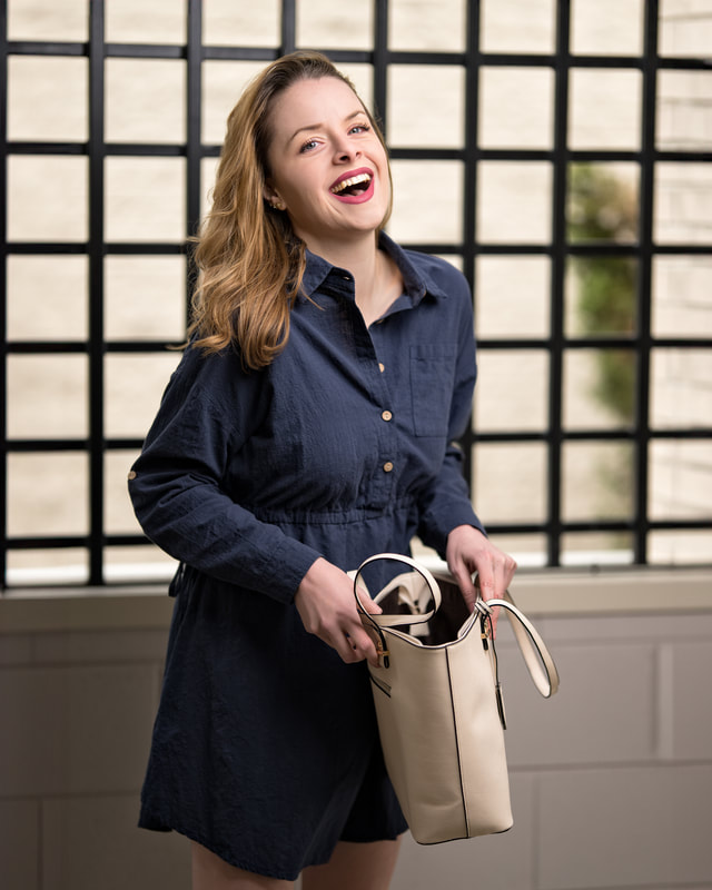 A photo of Maddie wearing a navy blue dress, laughing toward the camera, while holding open a beige colored purse. There is a black grate in the background and some concrete walls.