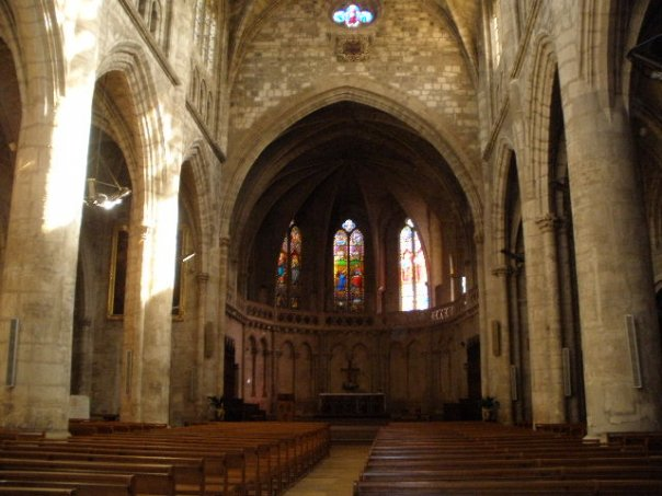 A photo of the inside of a dark cathedral with stained glass windows by the altar and flecks of sun on the columns.