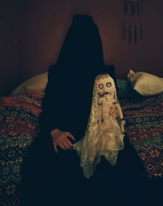 A spooky image of Maddie dressed in an all-black victorian mourning dress with a veil that completely covers her head, sitting on a bed holding a spooky white doll