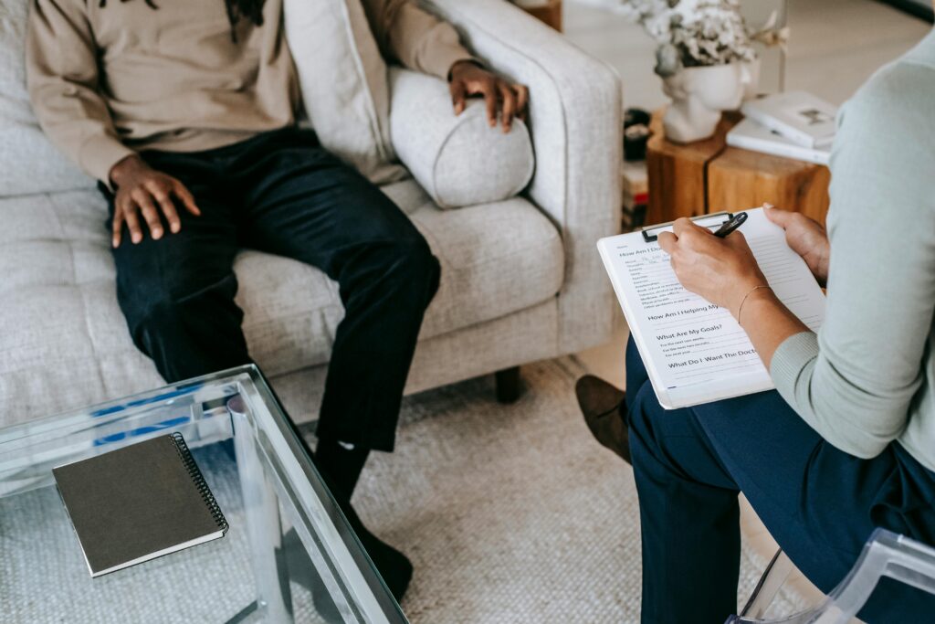 A person sitting on a couch speaking to another person who is writing on a clipboard