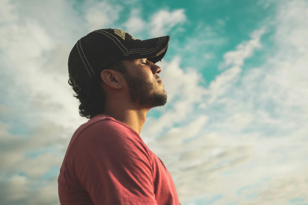 A man with a baseball hat outside with his eyes closed, appearing to be inhaling.