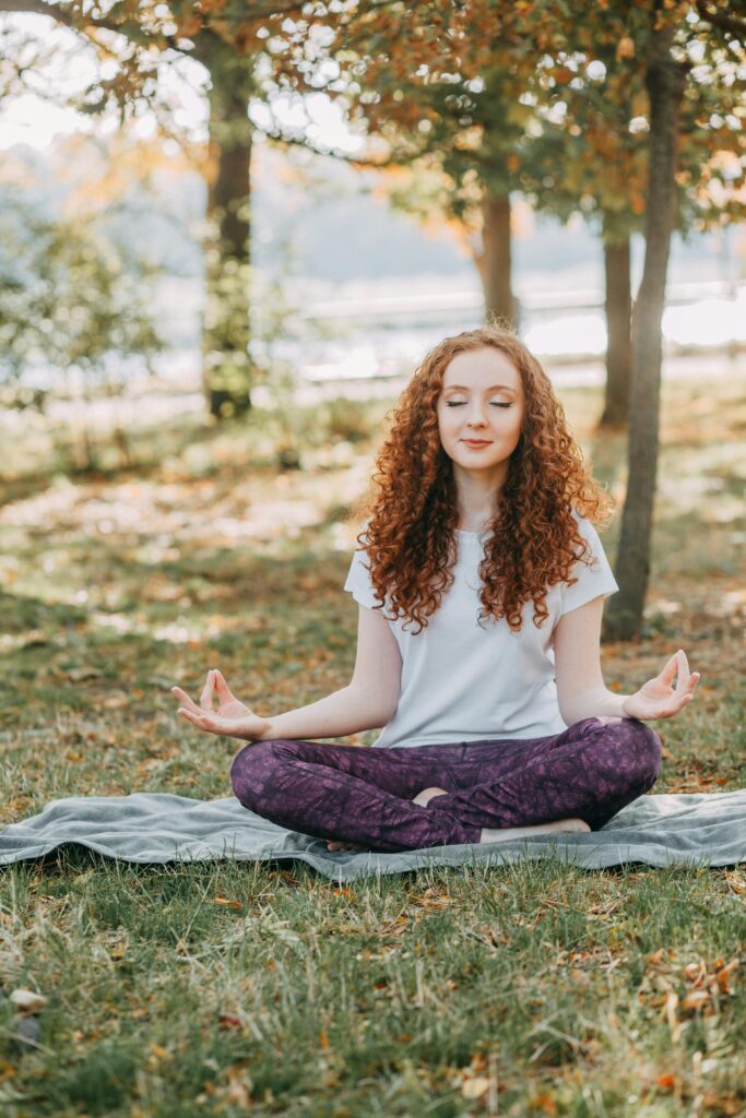 A woman sitting cross-legged on a blanket outside with her index fingers touching her middle fingers, hands resting on knees, and her eyes closed.