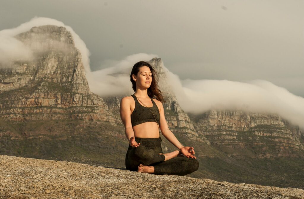 A stock photo of a woman in athletic apparel sitting on a foggy mountain cross-legged with her eyes closed.