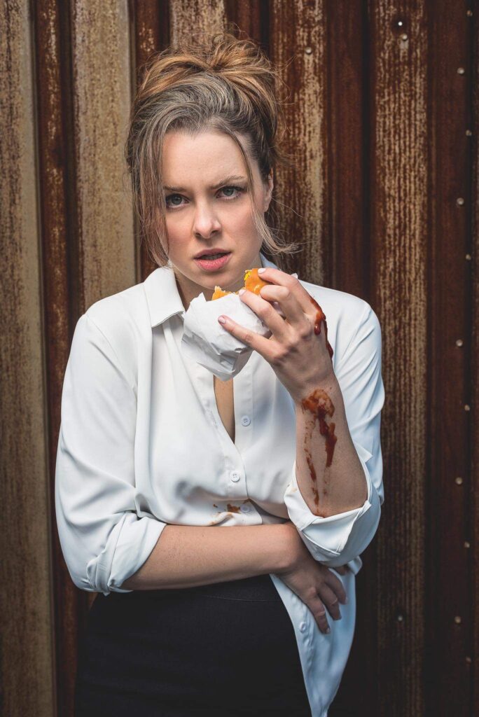 A photo of Maddie in a black pencil skirt and white blouse that is untucked, holding a cheeseburger in her left hand, with ketchup dripping down her left arm, looking toward the camera with a scowl on her face.