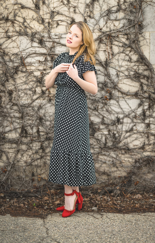 A photo of Maddie in a black and white polka dot short sleeved dress, standing in front of old vines on a concrete wall. She is wearing red shoes and is holding some of her hair in her hands.