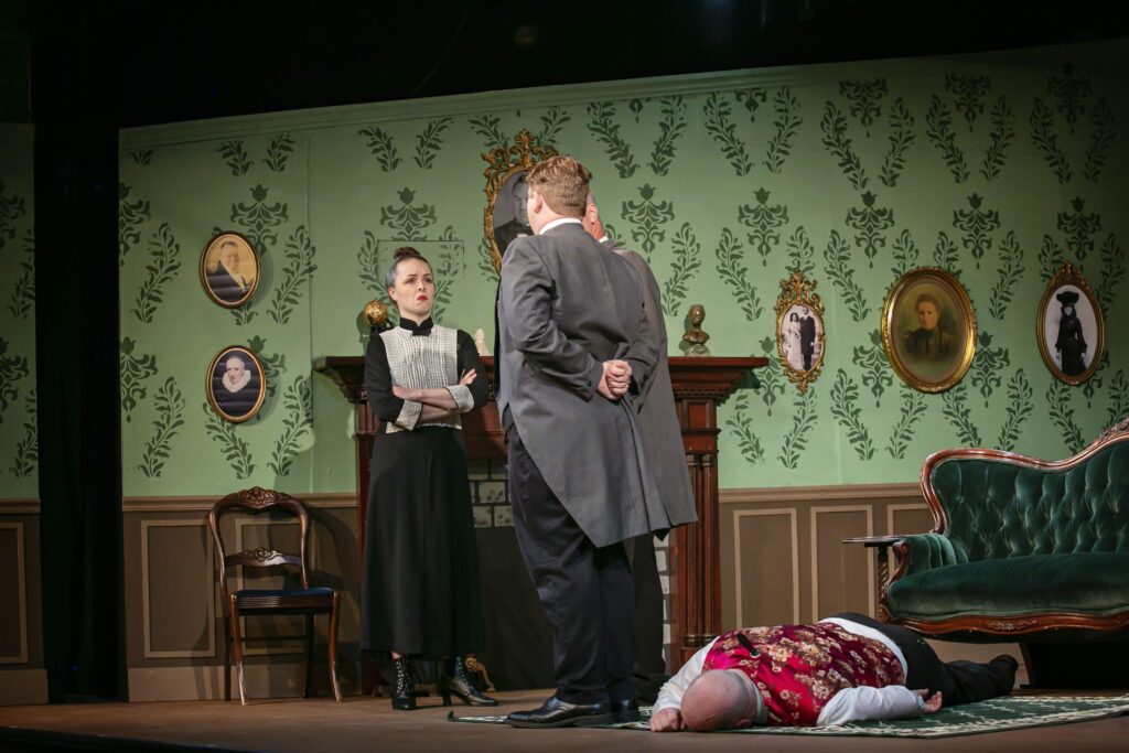 A photo of the theatrical production "Murder Comes to Uptight Abbey" with Maddie standing in front of a fireplace of a decorated wall with many old photos, with Marc and Luke facing her and Alex on the ground. 