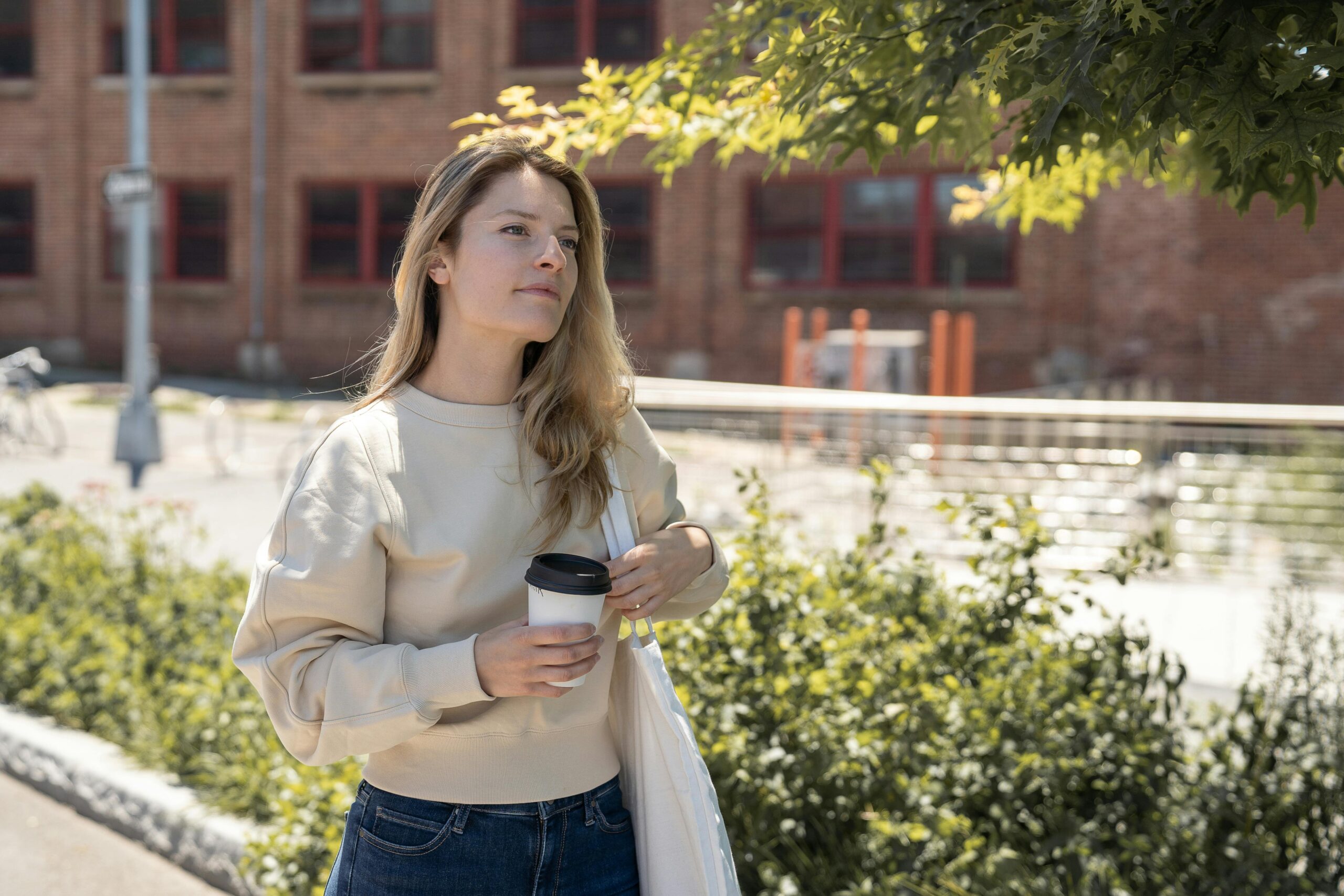 An image of a blonde woman carrying a cup of coffee outside near a bush with a light colored tote bag on her shoulder