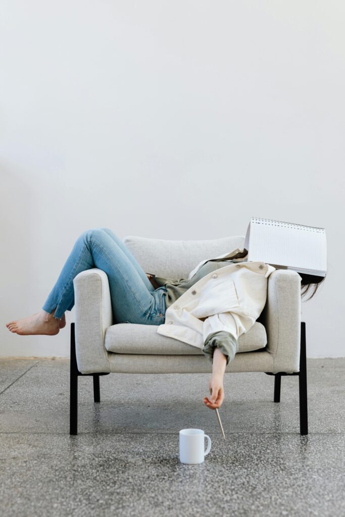 A photo of a woman lying on a couch with an open notebook over her face and a pencil in her hand, with a coffee cup sitting on the floor.