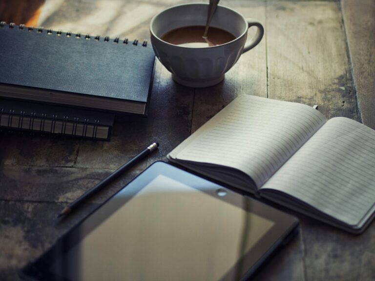 A photo of a coffee cup sitting on a table next to an open notebook with a pencil and other notebooks nearby