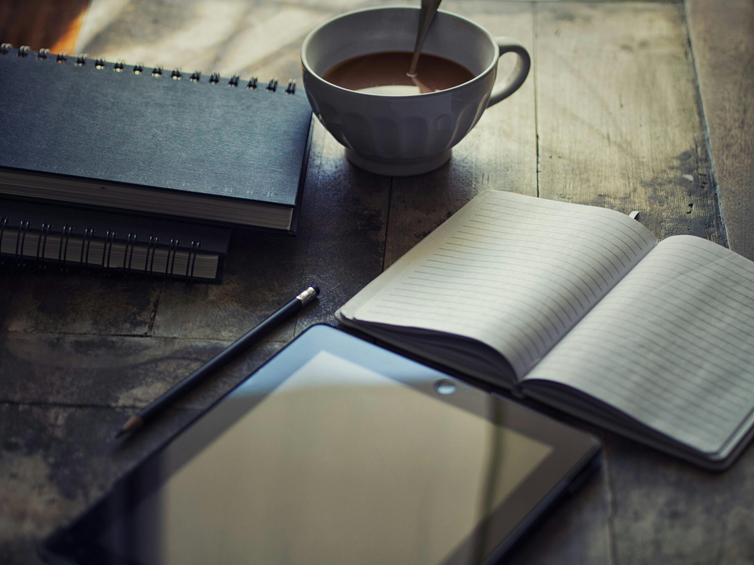 A photo of a coffee cup sitting on a table next to an open notebook with a pencil and other notebooks nearby