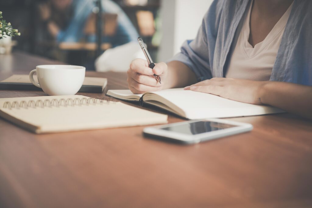 A photo of a woman holding a pen with an open notebook in front of her and a tablet with a coffee cup sitting on a desk.