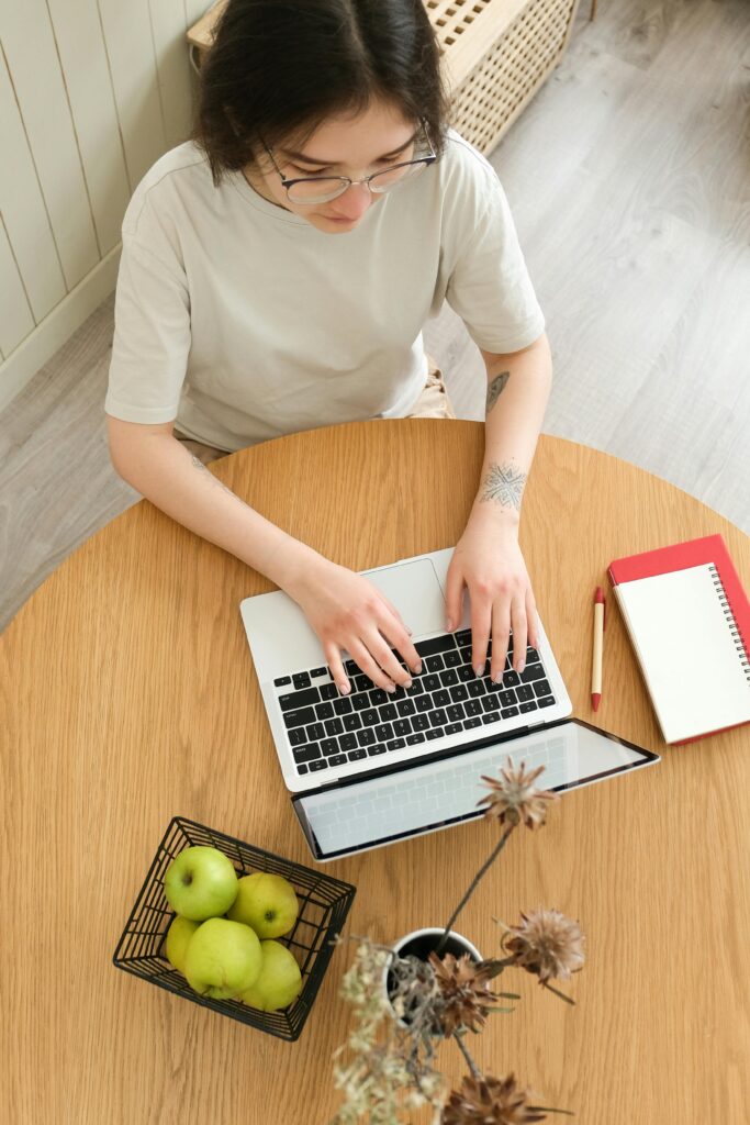 A photo of a woman with glasses working at a table on a laptop with a notebook and pencil next to her, and a basket of green apples nearby.